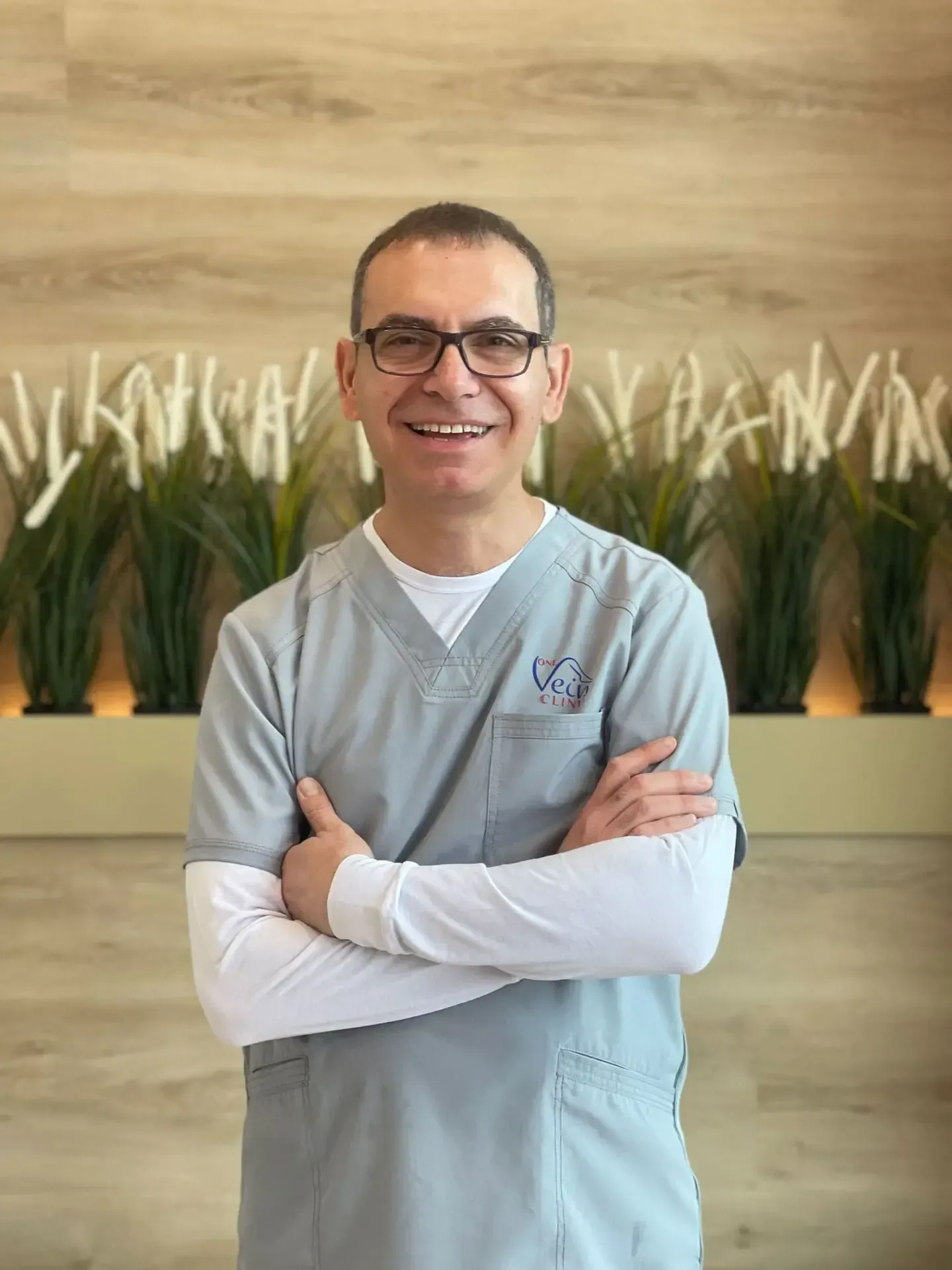 A man in a scrub top and glasses is posing for a picture with his arms crossed.