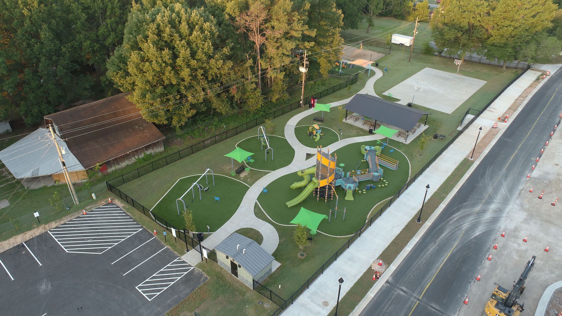Aerial view of a playground with various play structures, walkways, and a volleyball court, next to a building and road.