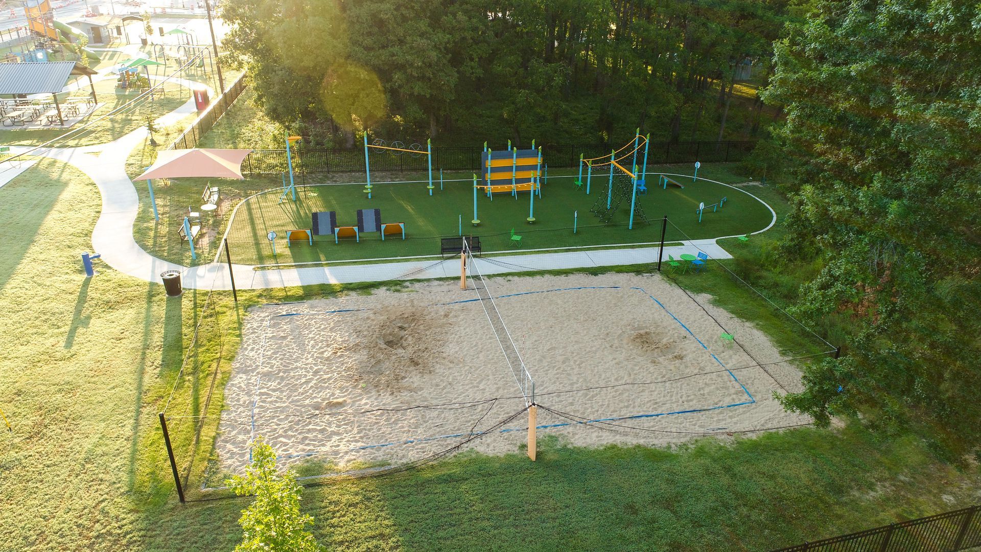 Park with a sand volleyball court and playground surrounded by grass and trees.