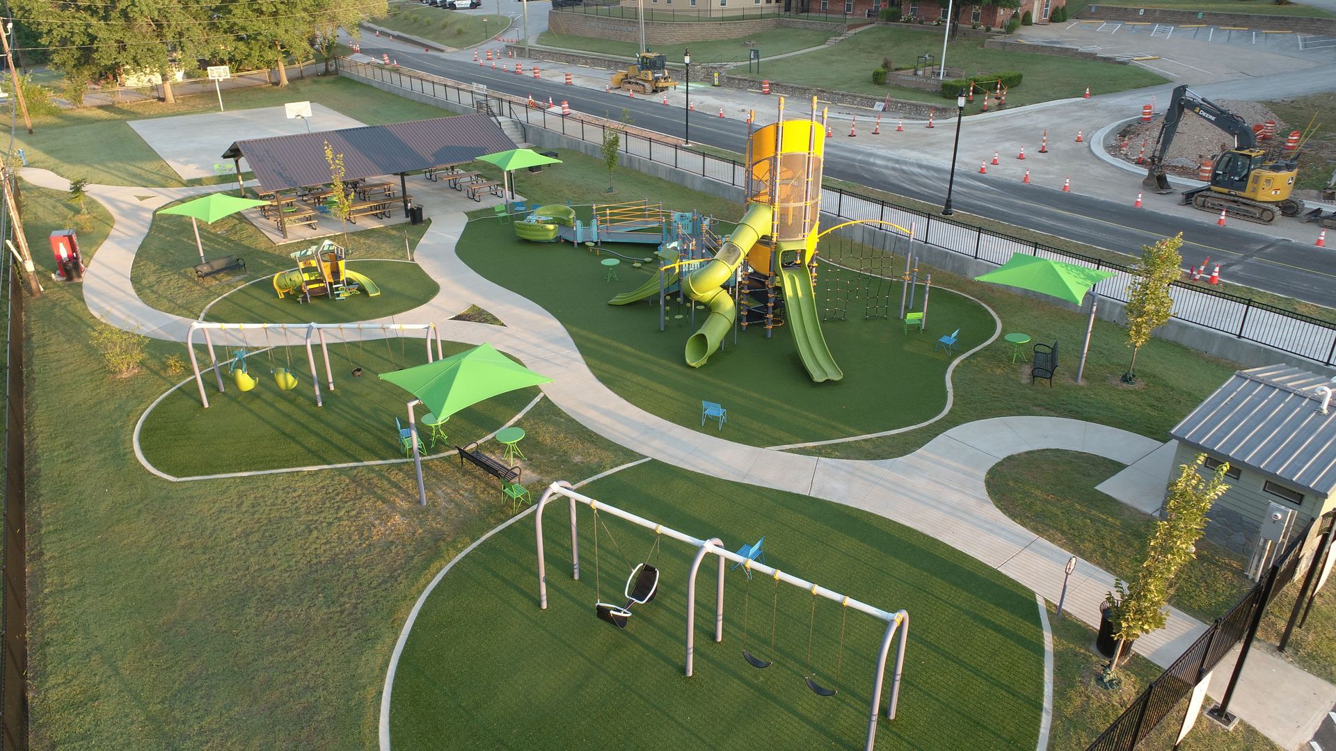 Overhead view of a modern playground with green turf, equipment, and paved walkways; green shade structures.