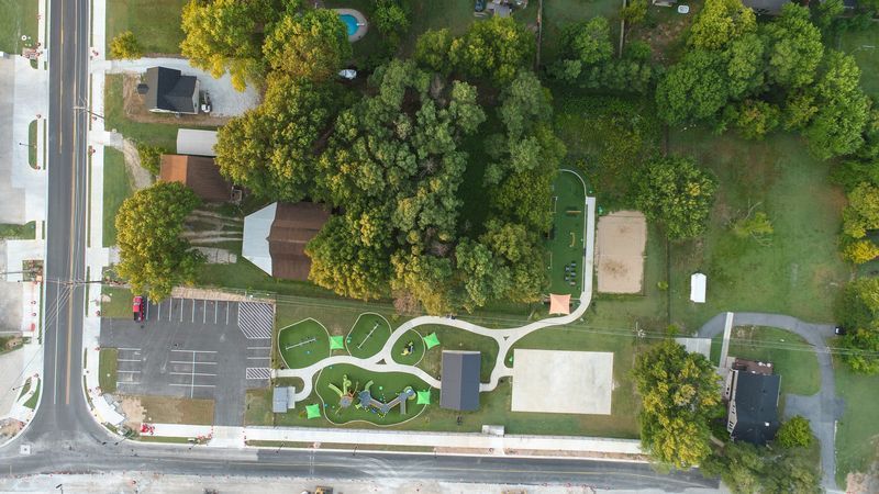 Aerial view of a park with playground equipment, a pathway, and surrounding trees and buildings.