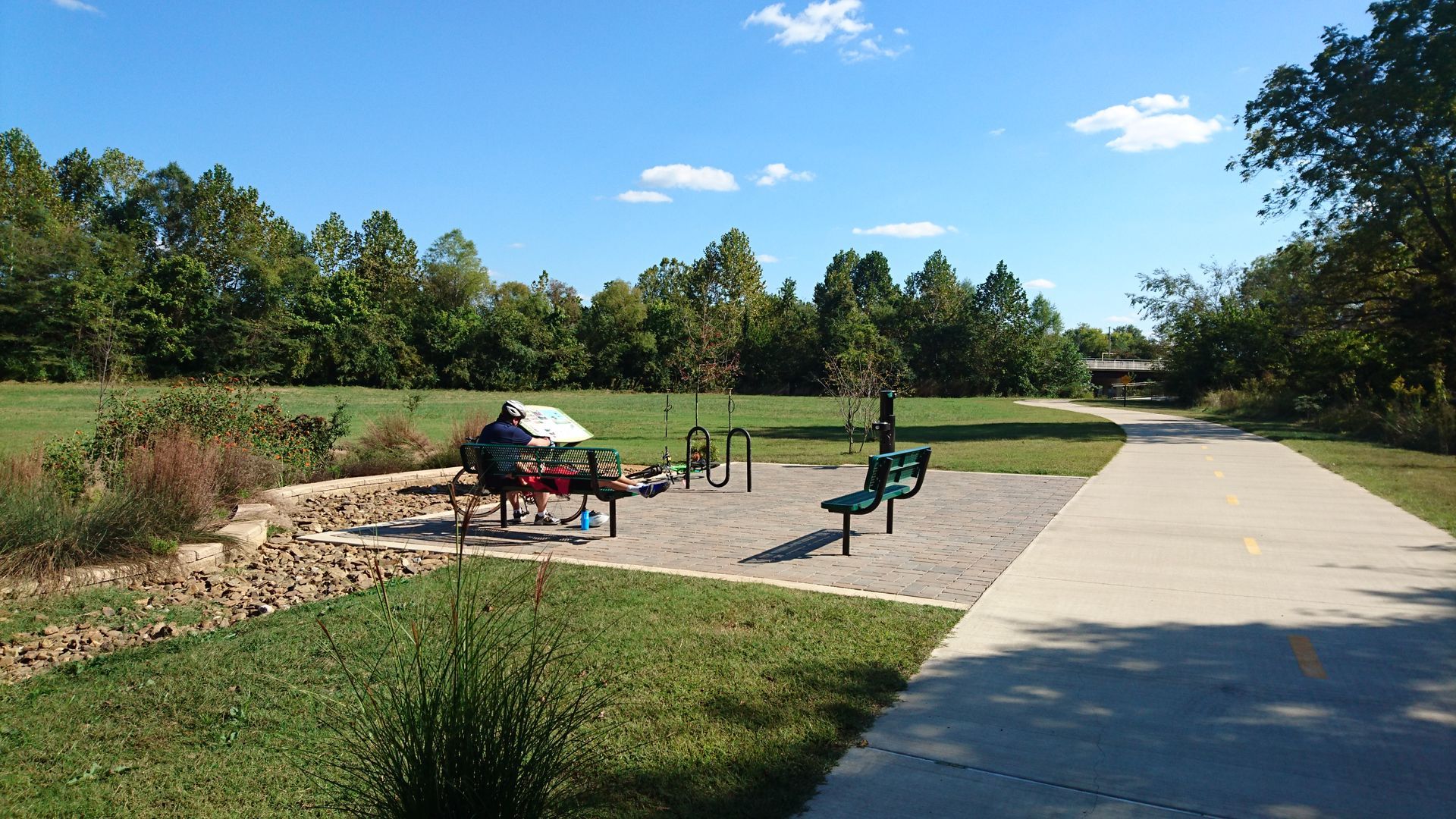 Park exercise equipment next to a paved path with a person using the equipment, surrounded by grass and trees.