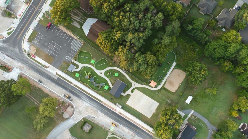 Aerial view of a park with playground, walking paths, sports court, trees, and parking lot.