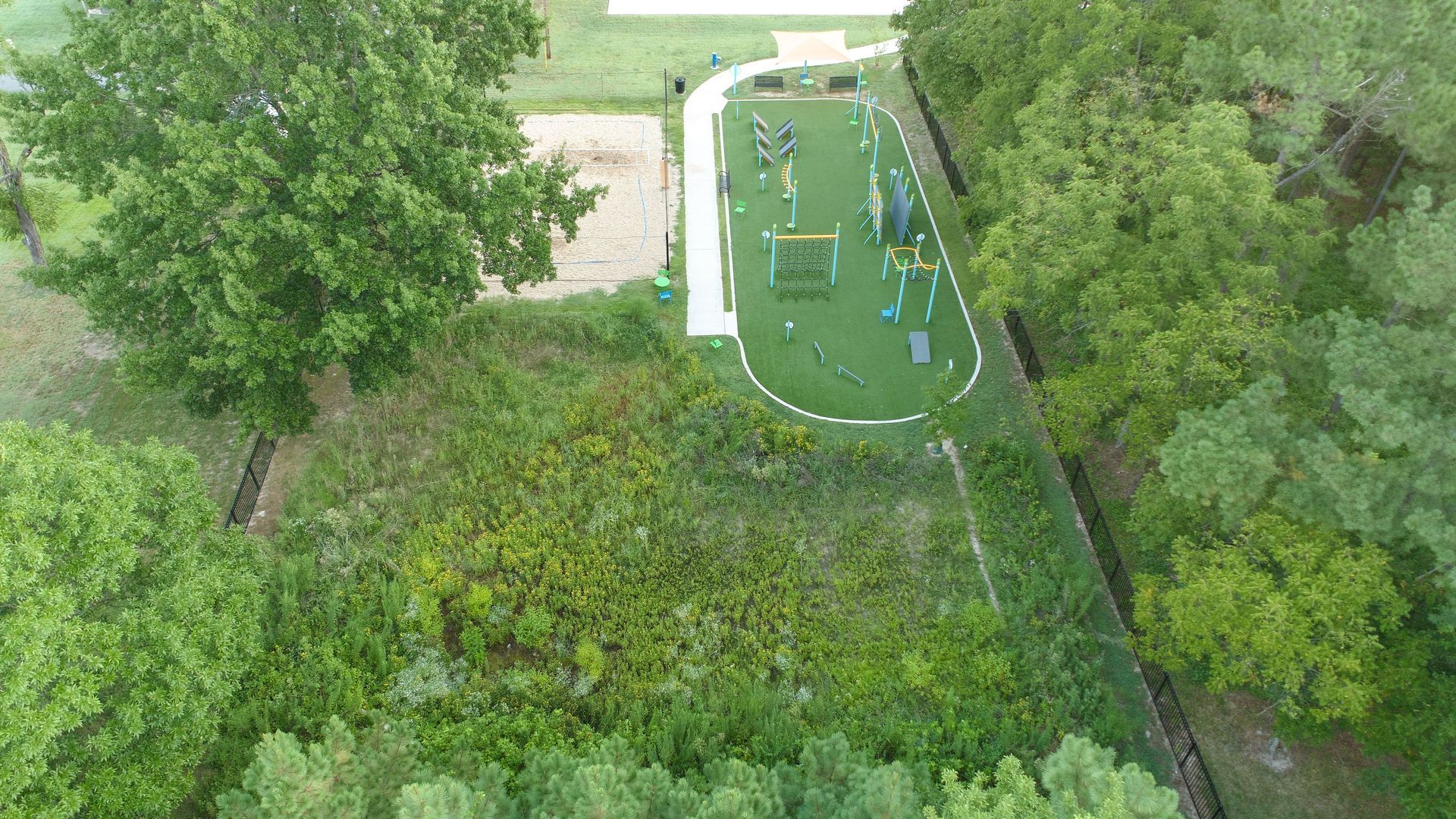 Aerial view of a playground with equipment on green turf, surrounded by trees and a long, white fence.