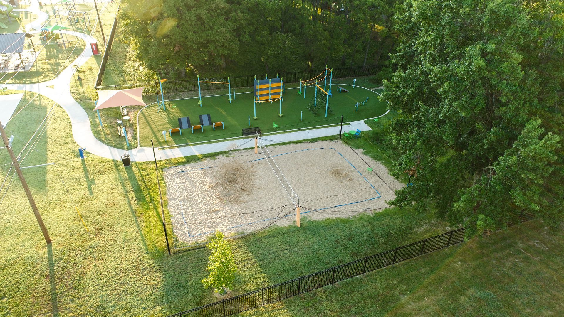 Aerial view of a recreational area with a sand volleyball court, playground, and walkway surrounded by trees.