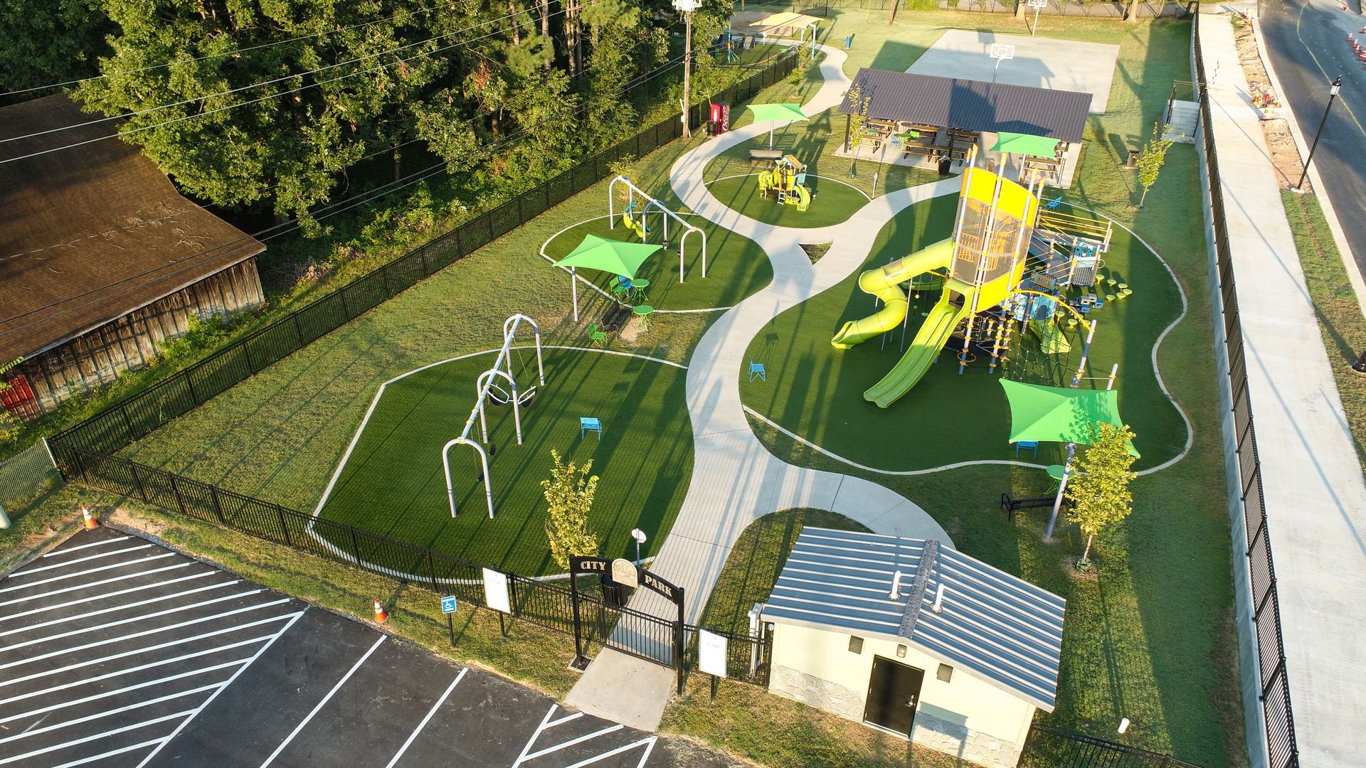Aerial view of a park with playground equipment, walking paths, and a small building, surrounded by grass.