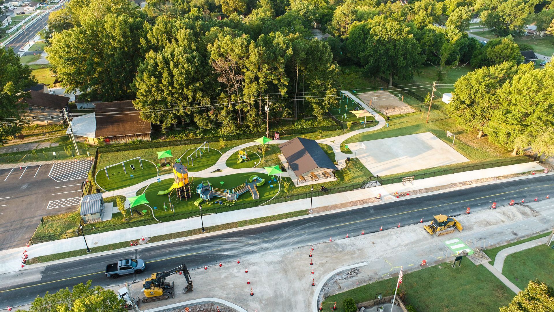 Aerial view of a park with playground equipment, a building, and a road under construction. Green trees surround the area.