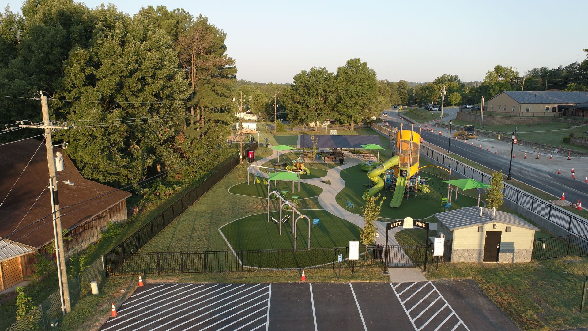Playground with various structures: slides, swings, and climbing features, enclosed by a fence and bordering a road.