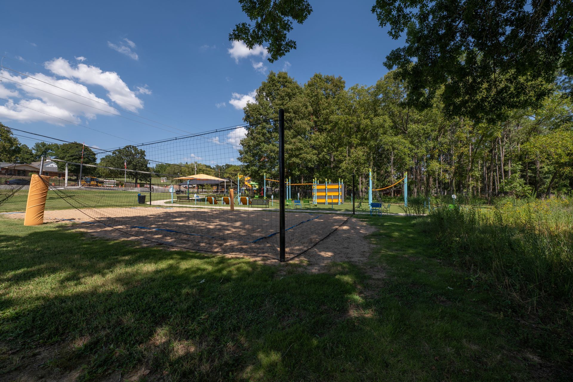 Volleyball court in a park on a sunny day. Net, poles, and structures are visible; trees in the background.