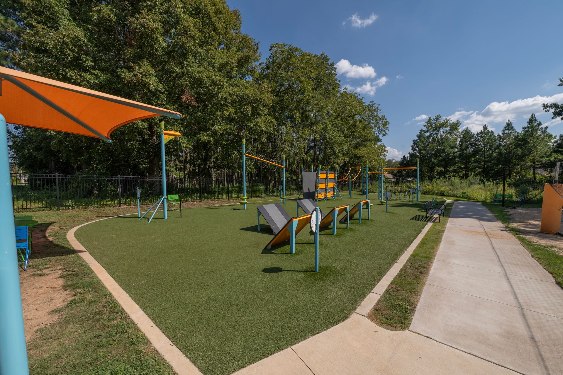 Outdoor fitness park with exercise equipment on artificial turf, under shade, with a walkway.