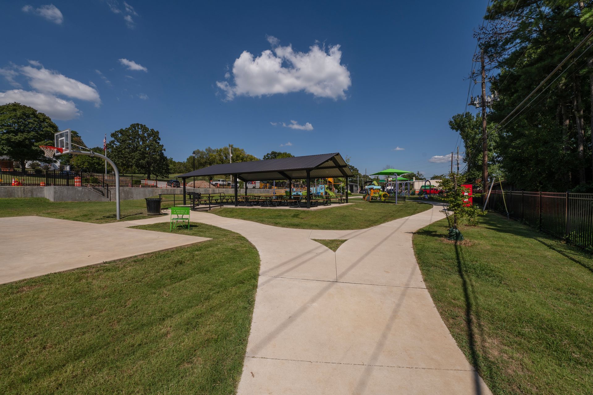Park with concrete paths, basketball court, playground, and covered picnic area under blue sky.
