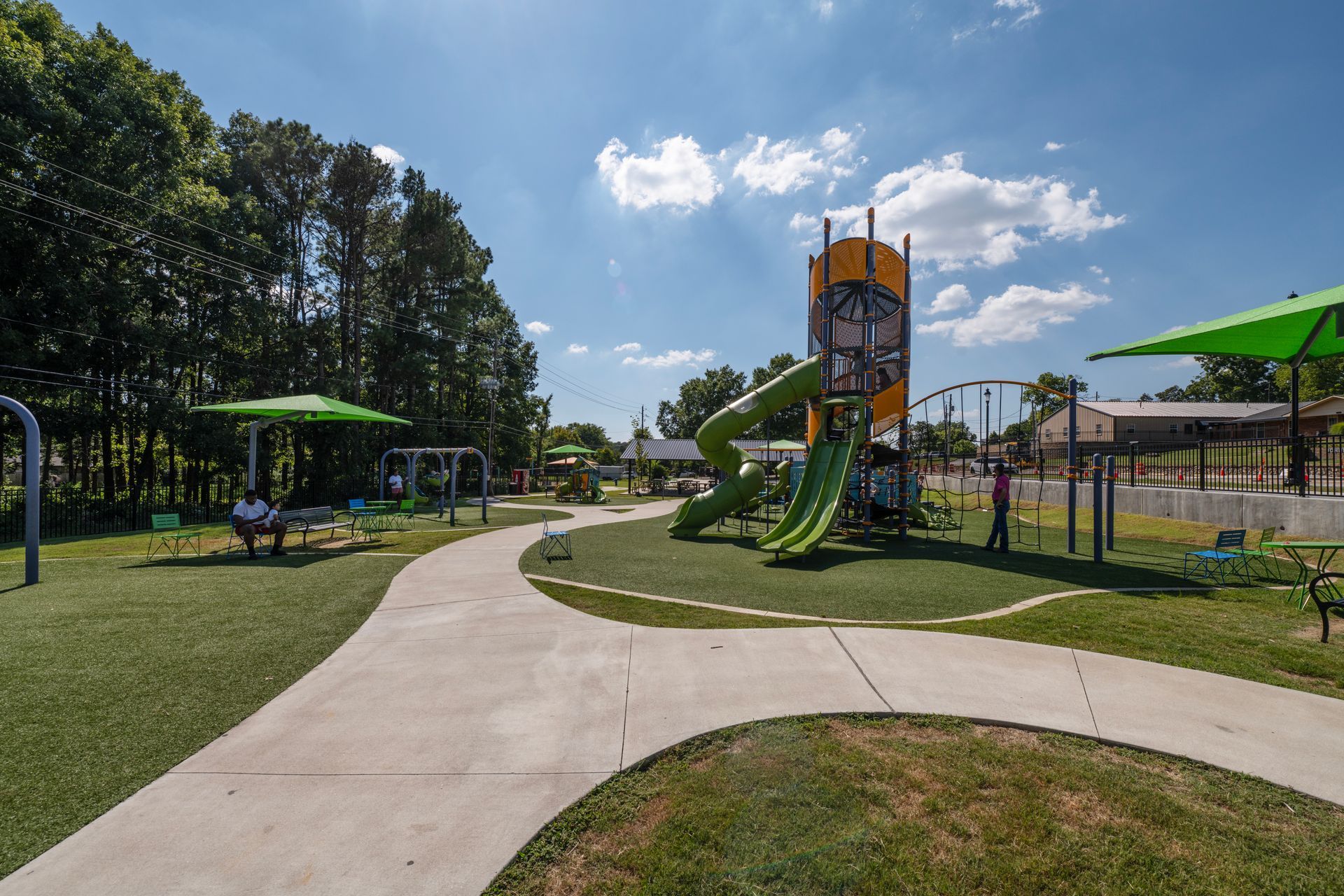 Playground with slides, swings, green turf, and concrete pathways under a blue sky.