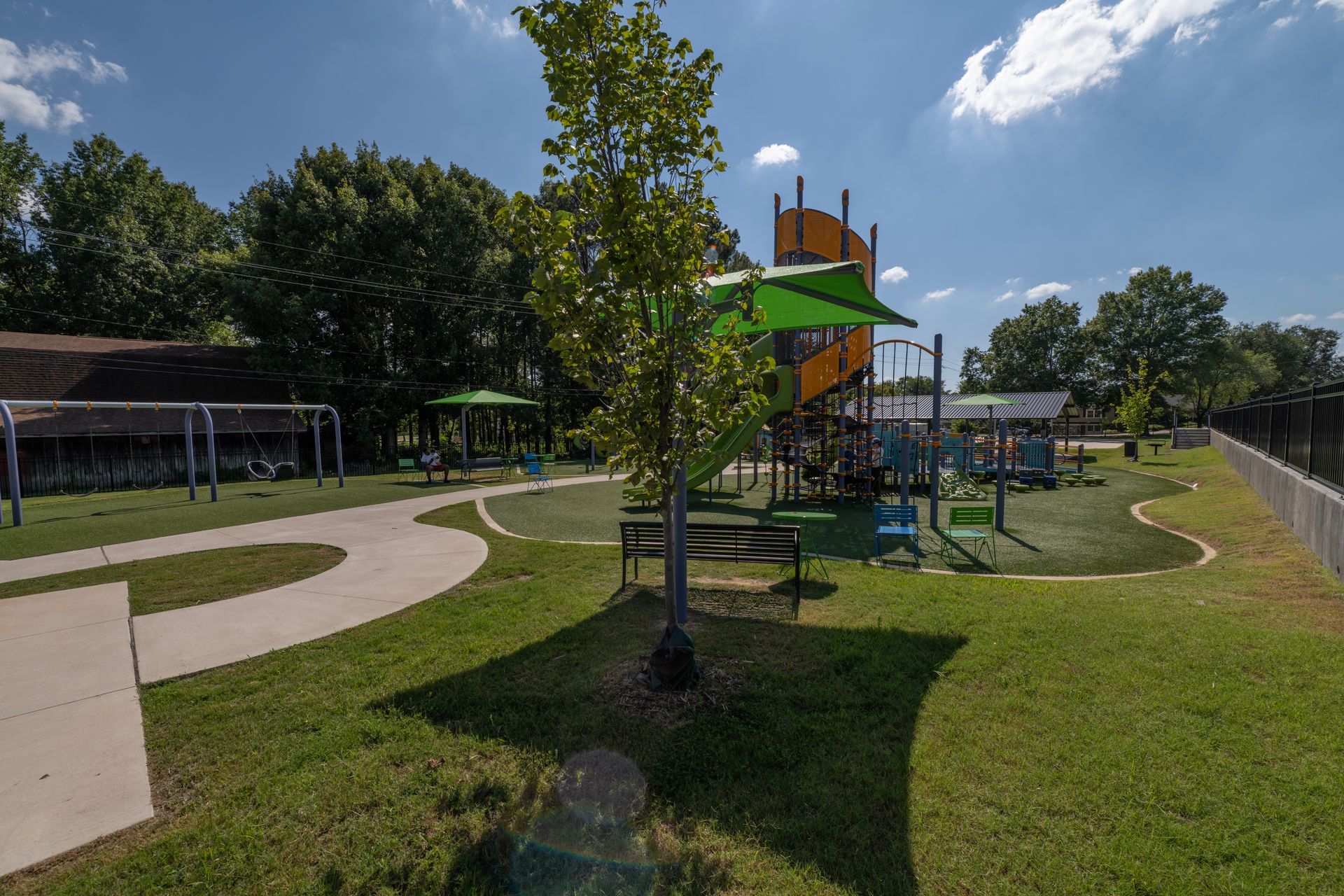 Playground with green grass, walkways, trees, and play structures under a bright blue sky.
