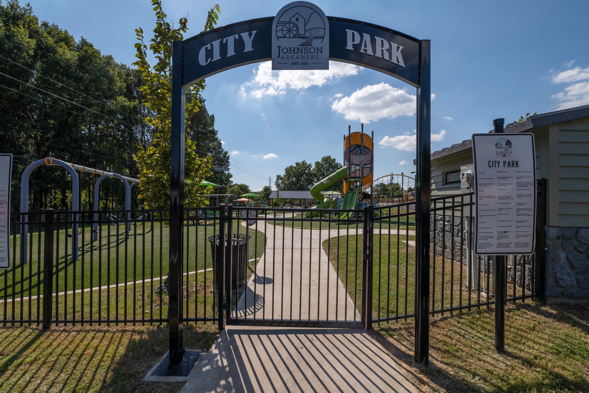 Black gate leading into City Park with a sign. Play structures are visible in the background under a blue sky.