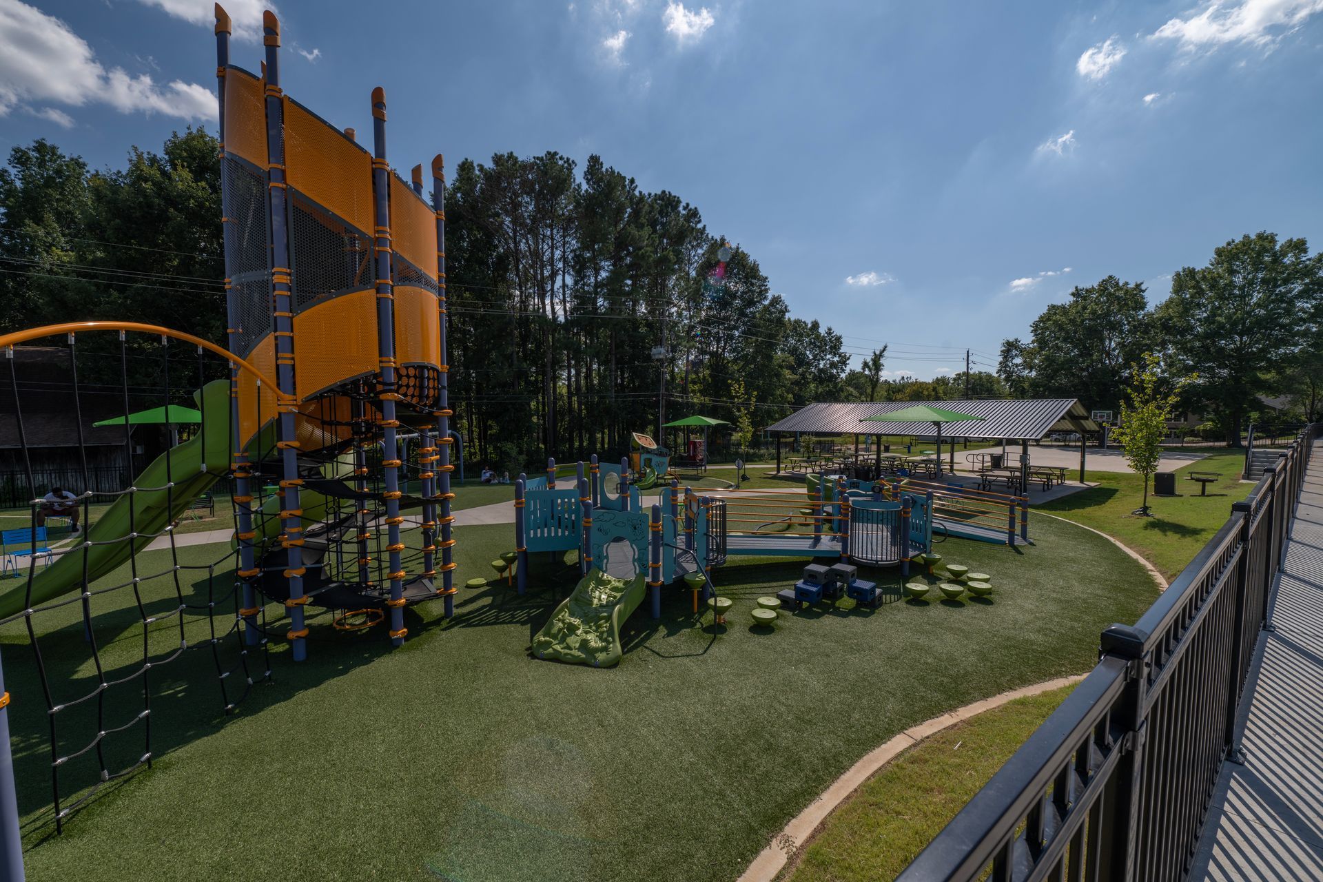 Playground with tall spiral slide, green turf, blue and yellow play structures, and a picnic shelter under a sunny sky.