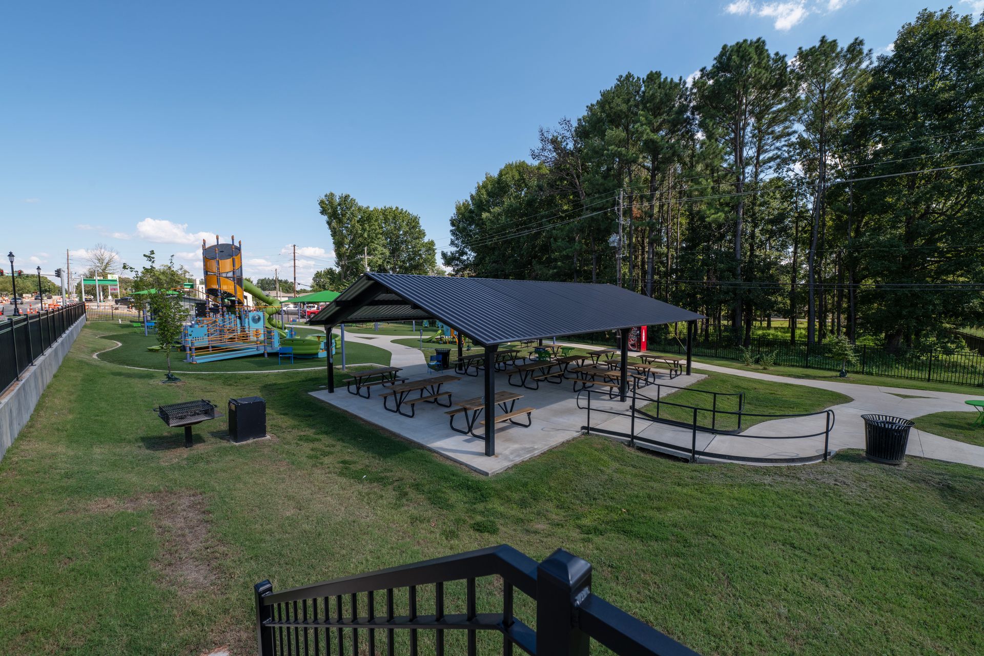 Park with picnic tables under a shelter, playground equipment, trees, and a black fence under a blue sky.