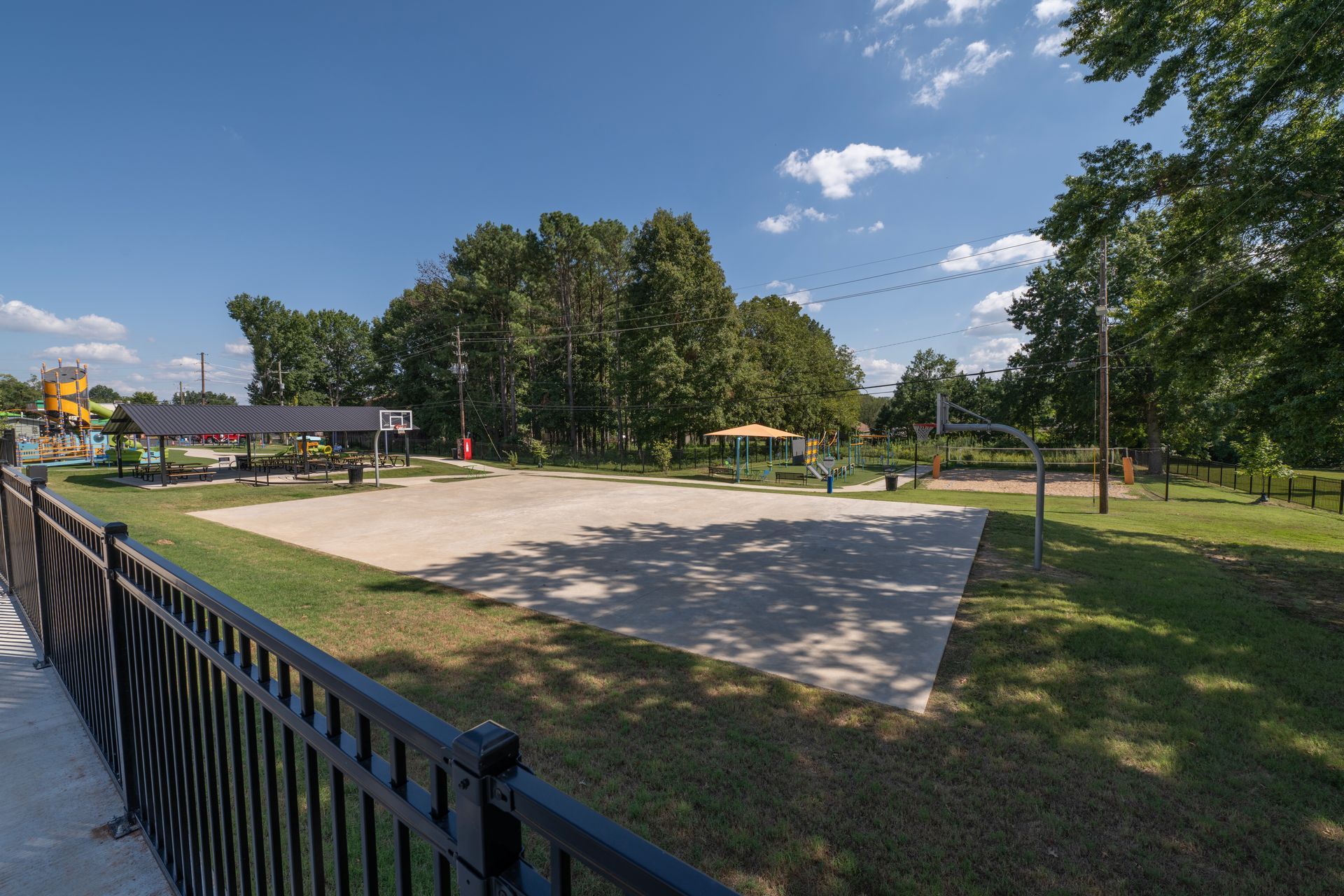Sandy volleyball court in a park with trees and a black fence. Blue sky.