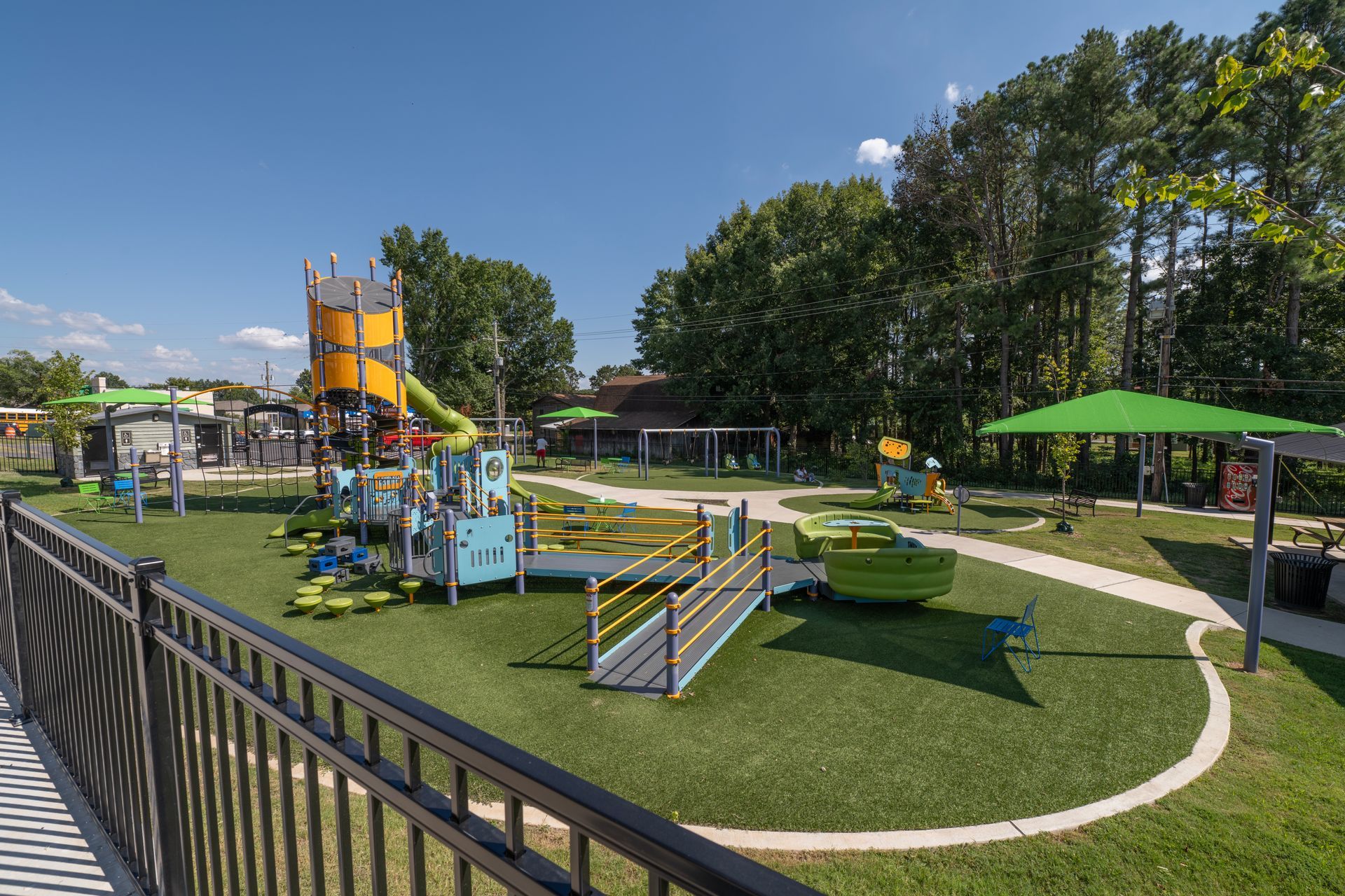 Playground with colorful play structures, green turf, sunny day, black fence.