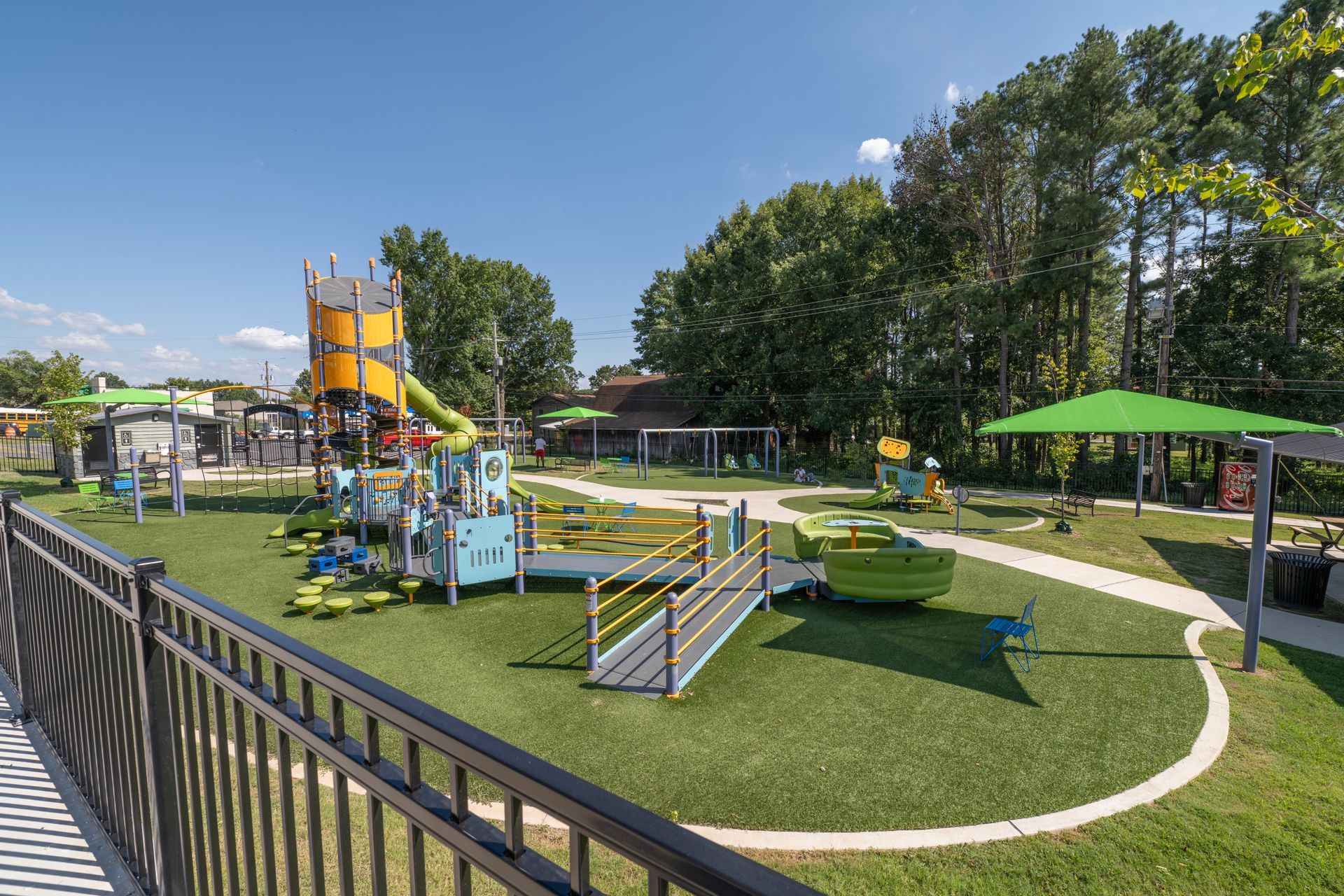 Playground with colorful equipment, green turf, trees, and covered seating areas.