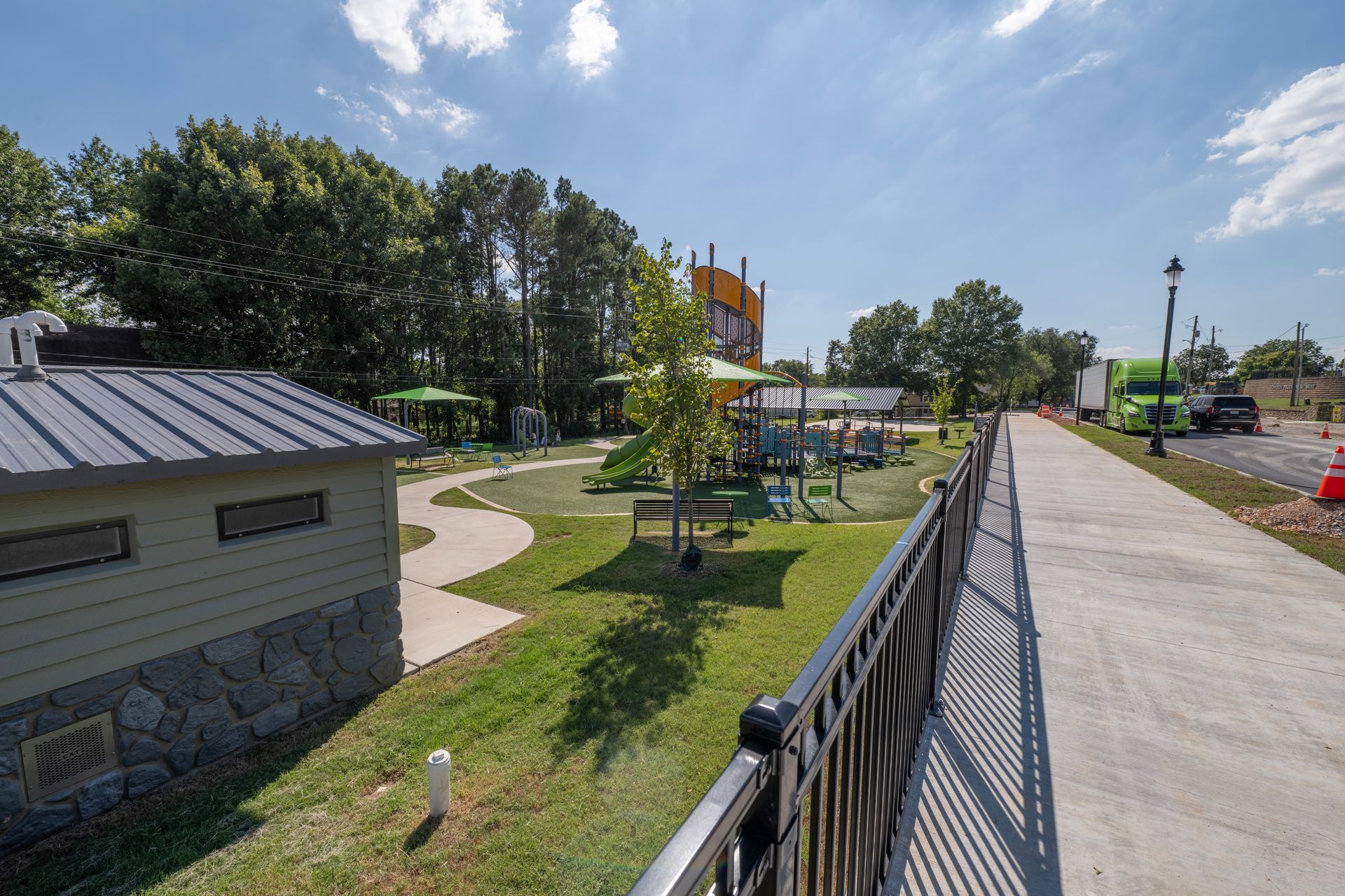 Playground with green grass, trees, and play structures. Sidewalk and restroom building in foreground. Bright, sunny day.