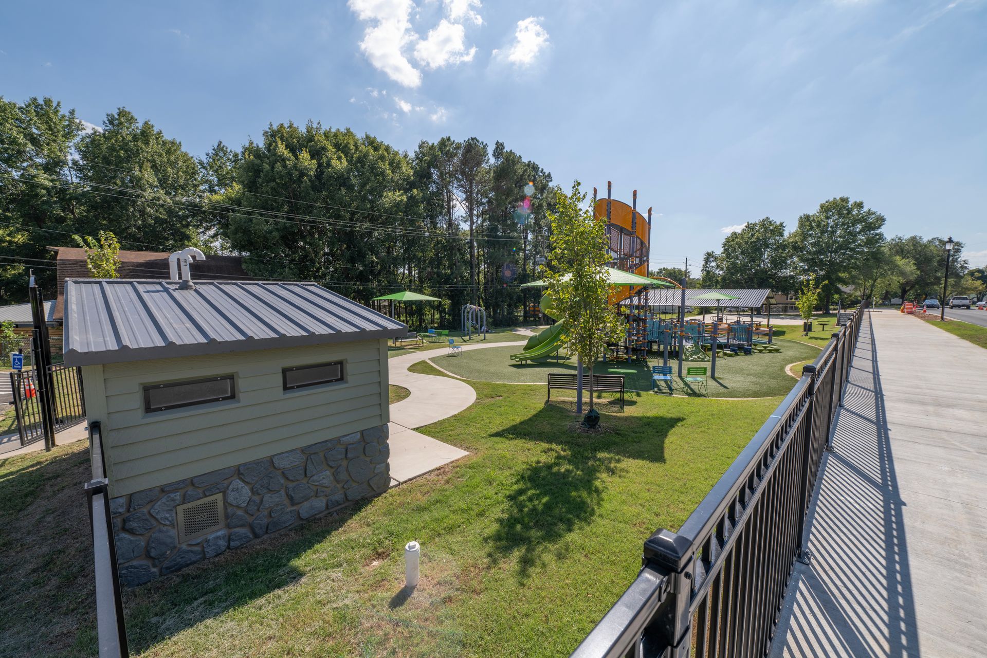 Park with playground equipment and a small building with a metal roof, sunny day.