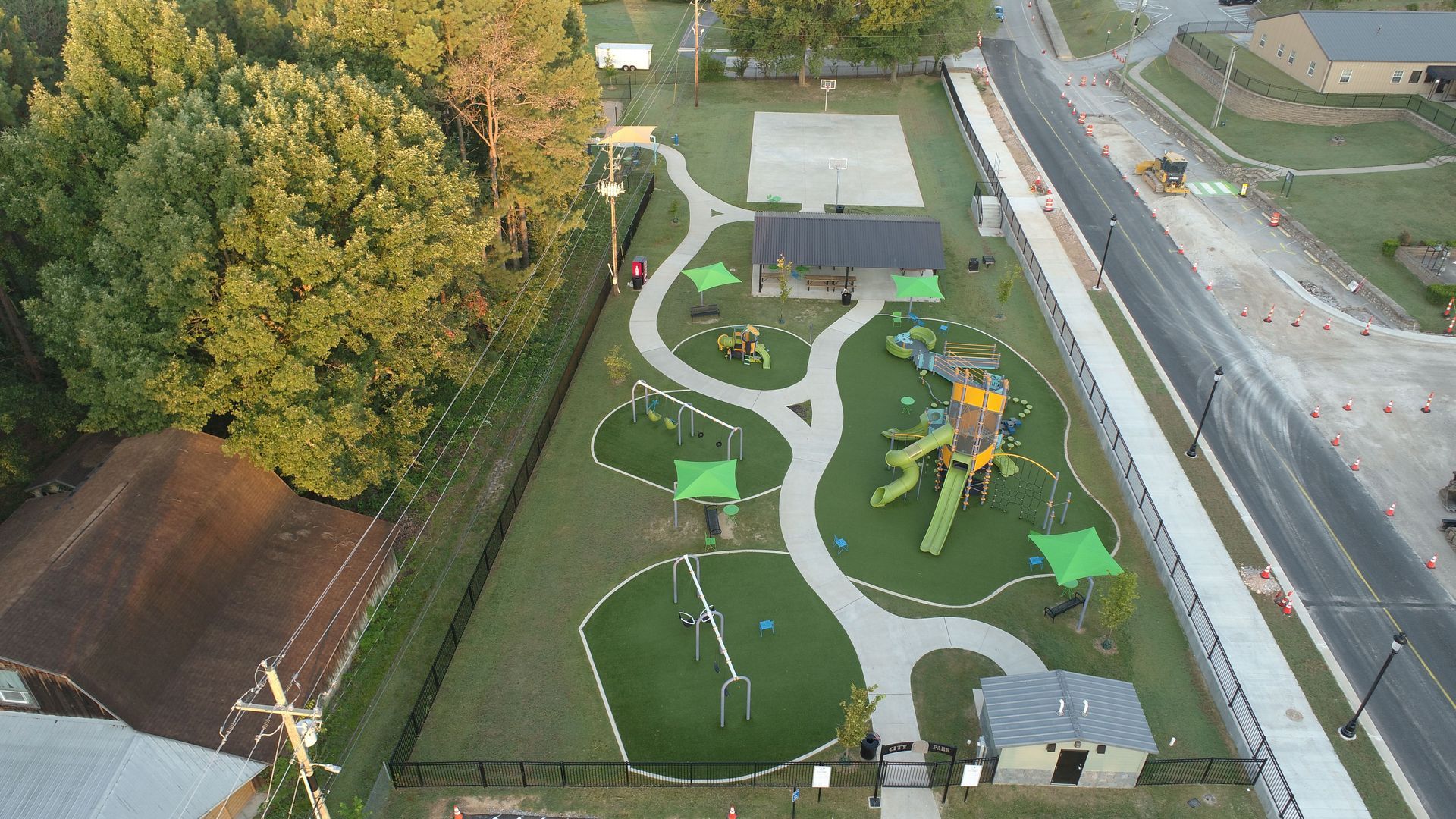 Aerial view of a park with playground equipment, walking paths, shelters, and a basketball court, next to a road.
