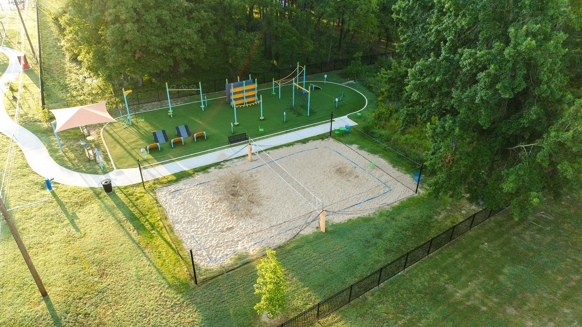 Aerial view of park with sand volleyball court, playground, path, gazebo, and trees.