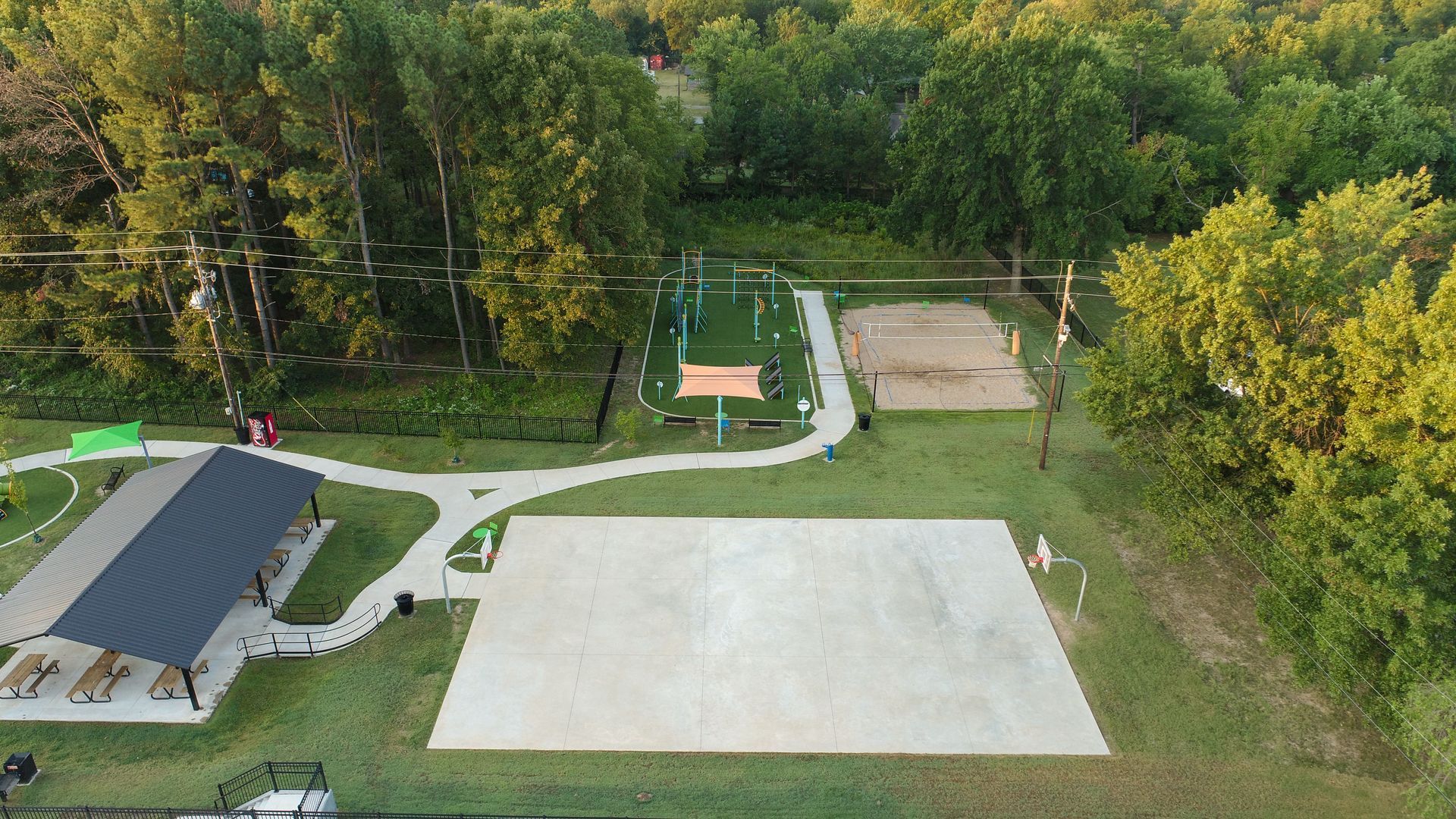 Aerial view of a park with a concrete area, playground, sand volleyball court, pavilion, and trees.