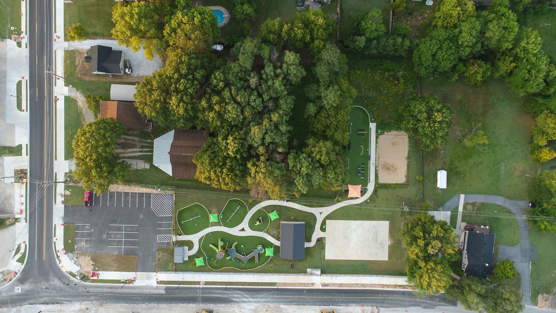Overhead view of a park with playground, trees, buildings, parking lot, and streets.