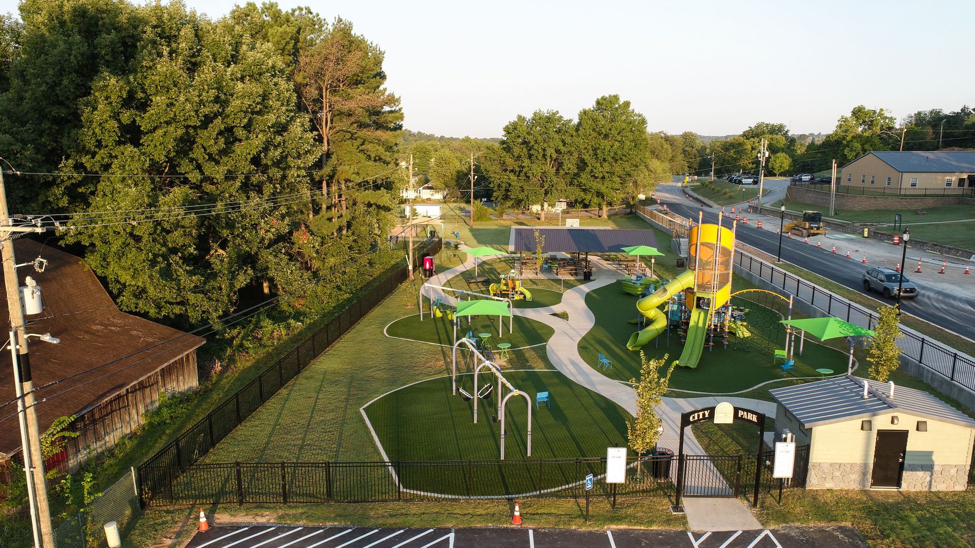 Aerial view of a brightly colored playground with green turf, equipment, trees, and a restroom building.