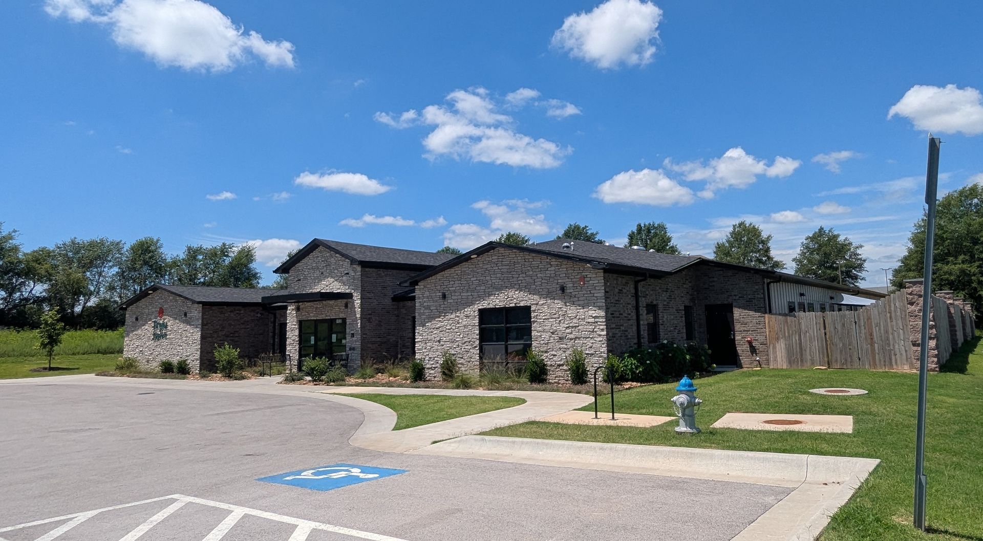 Stone building with dark roof under a blue sky, accessible parking space in front, grass and trees around.