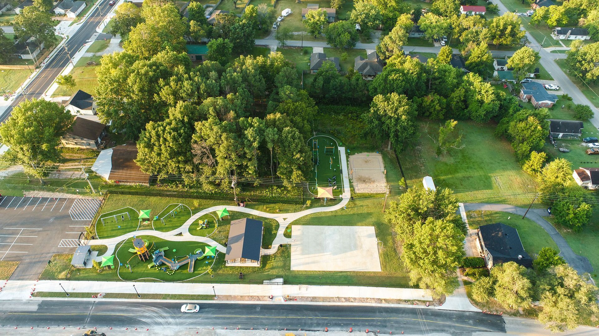 Aerial view of a park with playground equipment, a basketball court, and surrounding trees and houses.