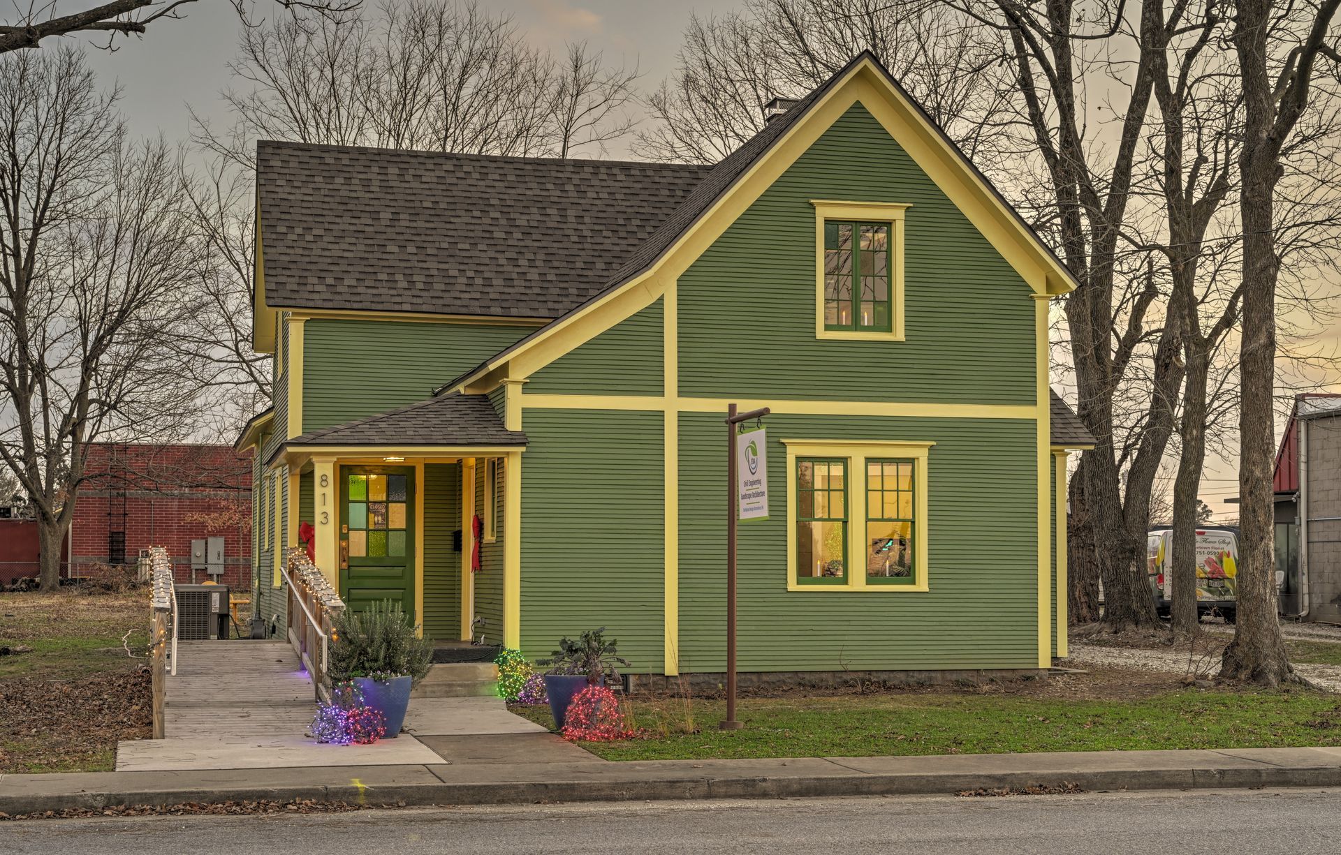 Green house with yellow trim, flowers in pots, trees in the background, and a sidewalk.