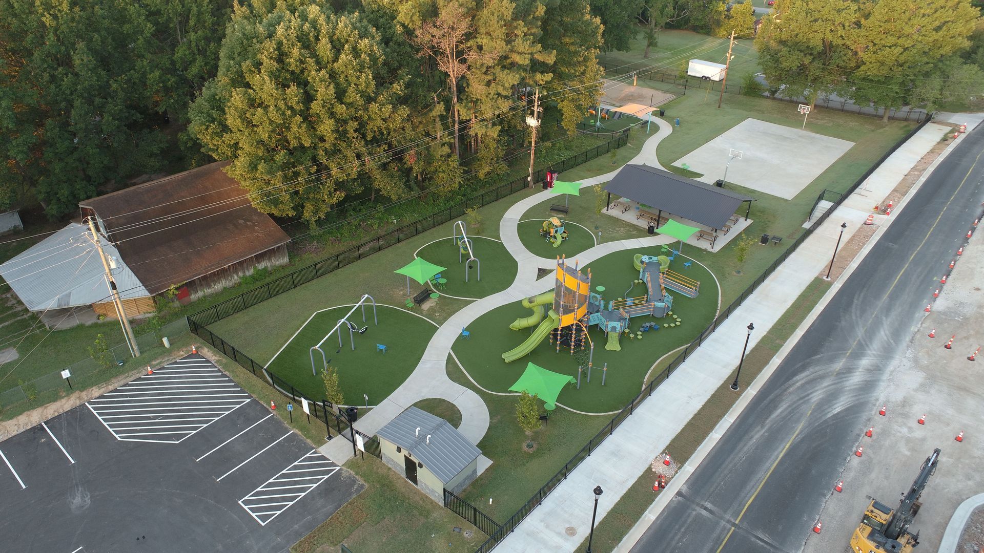 Aerial view of a playground with play structures, paths, and a covered shelter, next to a parking area and street.