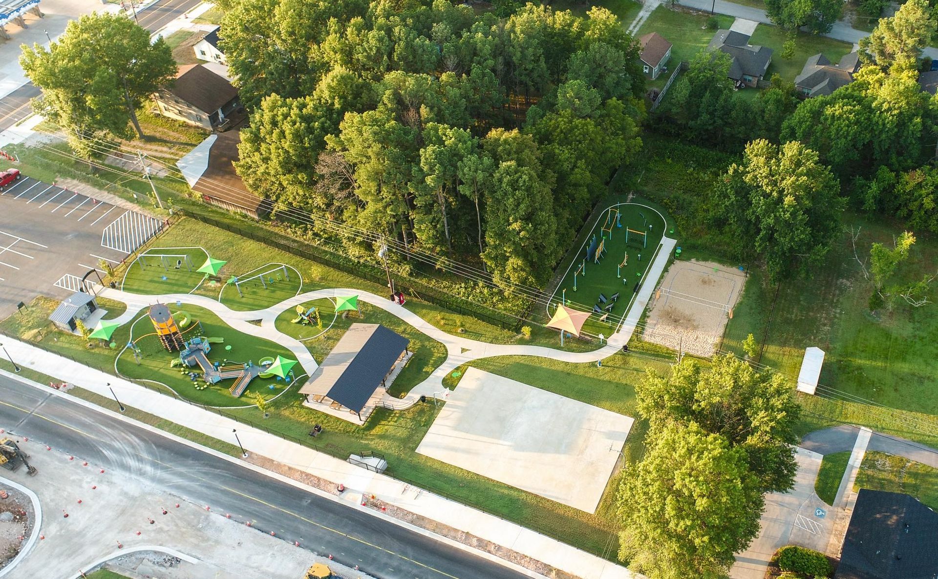 Aerial view of a park with playground, paths, sports area, and surrounding trees; sunny day.