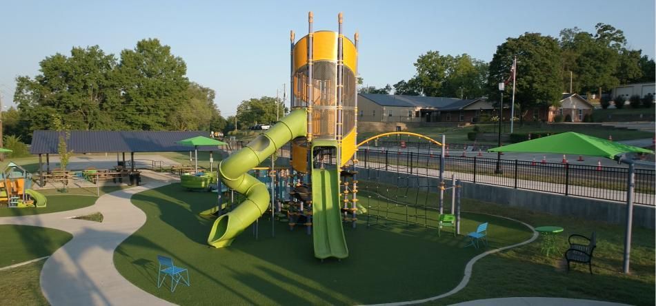 Playground with a tall yellow and green slide tower, green turf, and trees.
