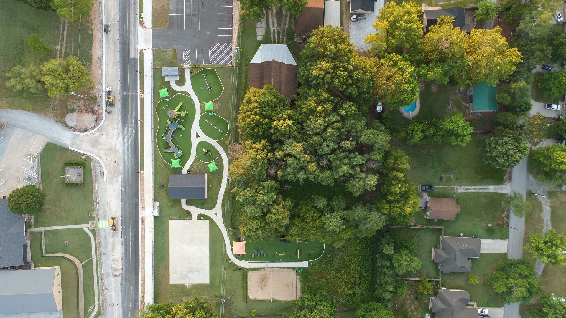 Aerial view of a park with playground equipment, trees, paths, and adjacent road and buildings.