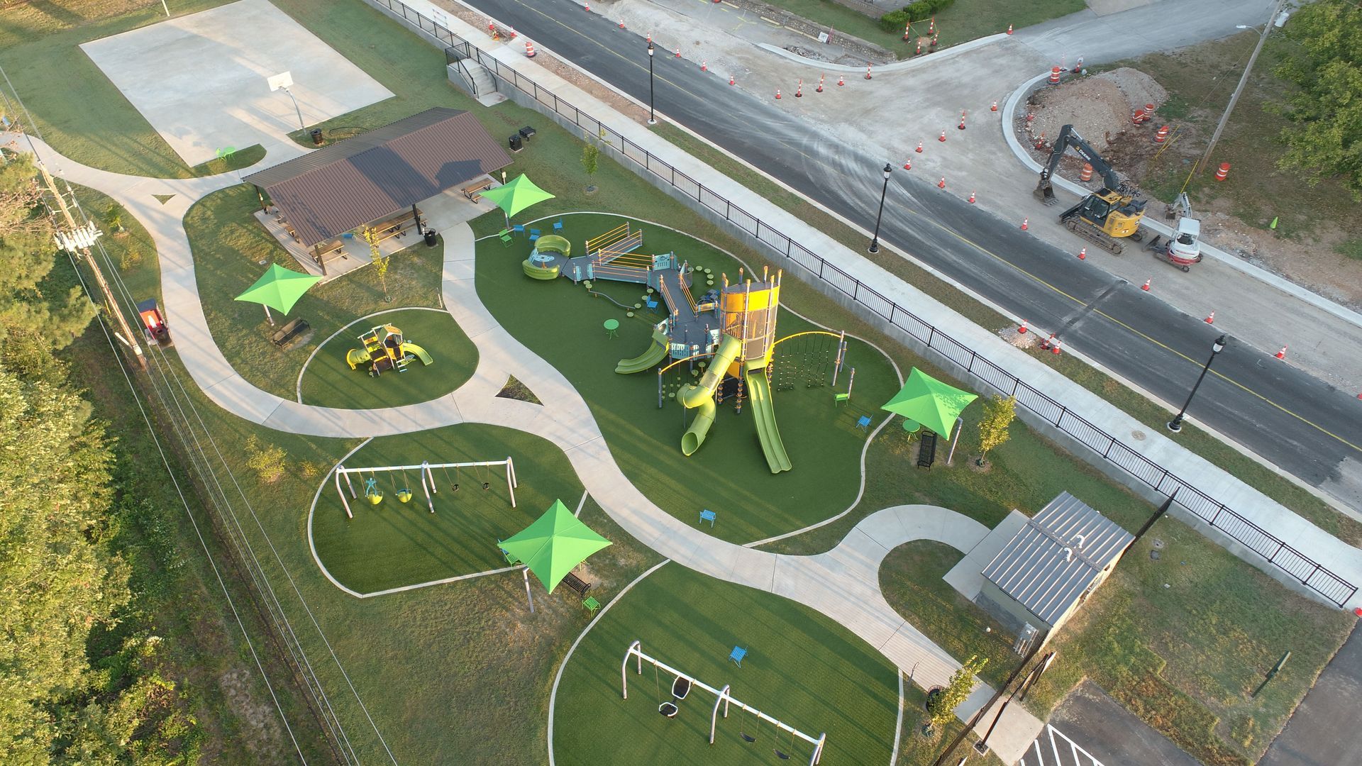 Aerial view of a playground with green turf, play structures, paths, and shade structures, near a road.