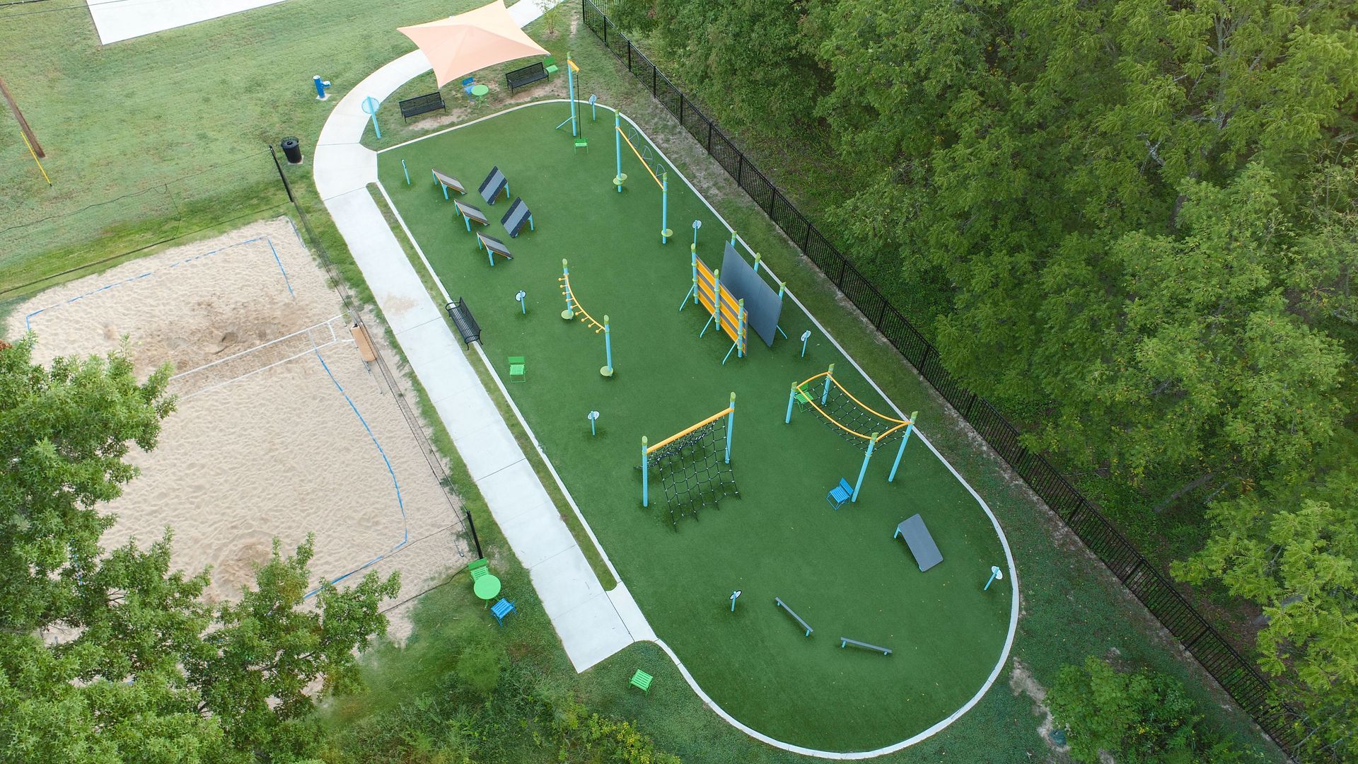Aerial view of a dog park with agility equipment, artificial turf, sand pit, and surrounding trees.
