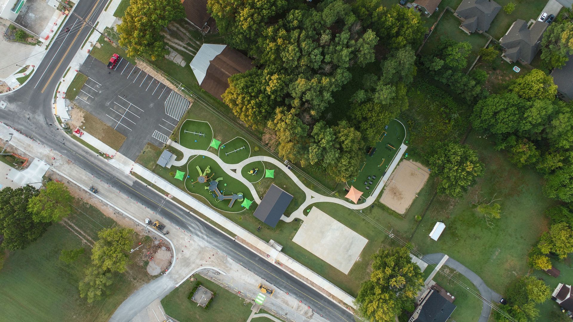 Aerial view of a park with playground equipment, walking paths, and a parking lot next to a road and trees.