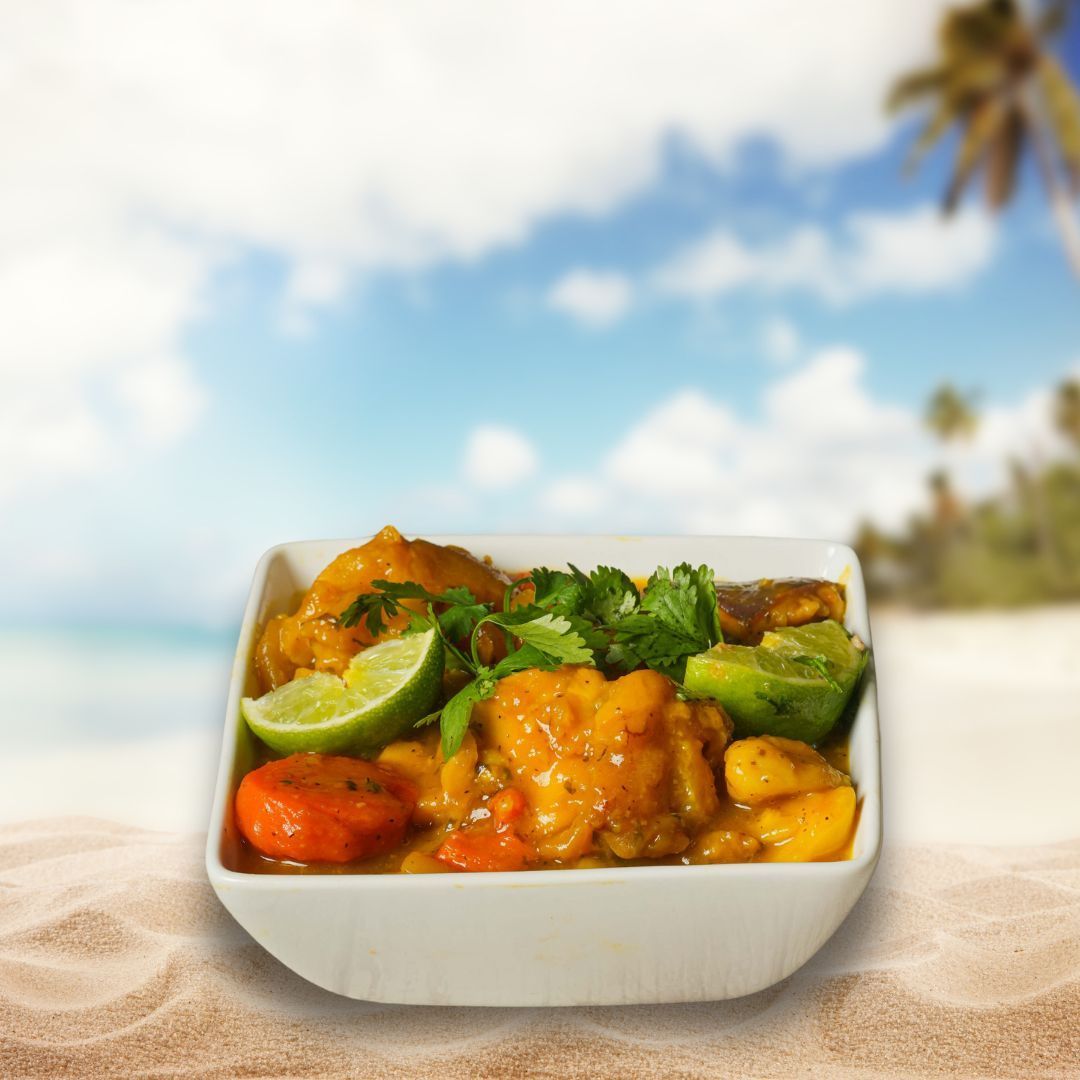 A white bowl of orange curry with cilantro and lime wedge on a sandy beach with a tropical background.