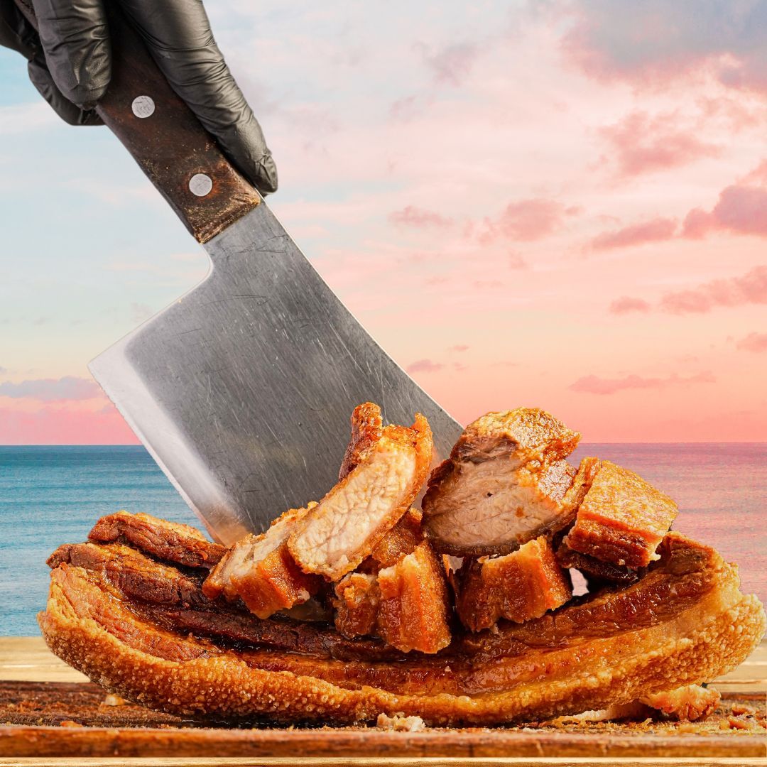 A cleaver chops crispy pork belly on a wooden board. The hand holding the cleaver wears a black glove