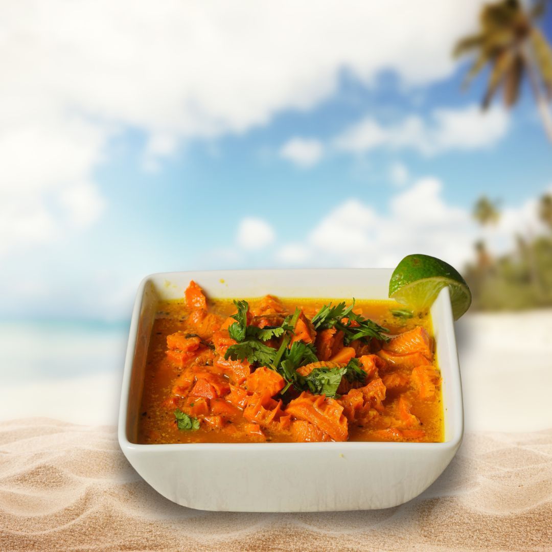 A white bowl of orange curry with cilantro and lime wedge on a sandy beach with a tropical background.