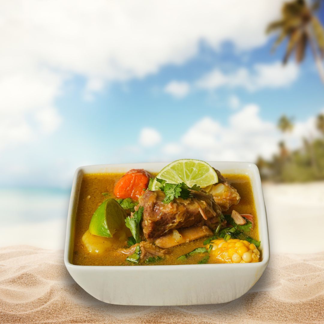 A bowl of Caribbean stew with meat, vegetables, and a lime wedge on a sandy beach with a blurred ocean and palm trees.