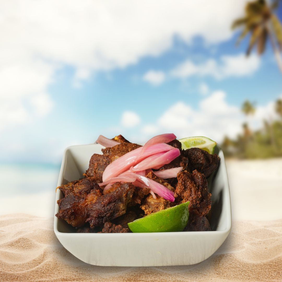 Bowl of jerk pork with pickled onions and lime, on a sandy beach with ocean and palm trees in the background.
