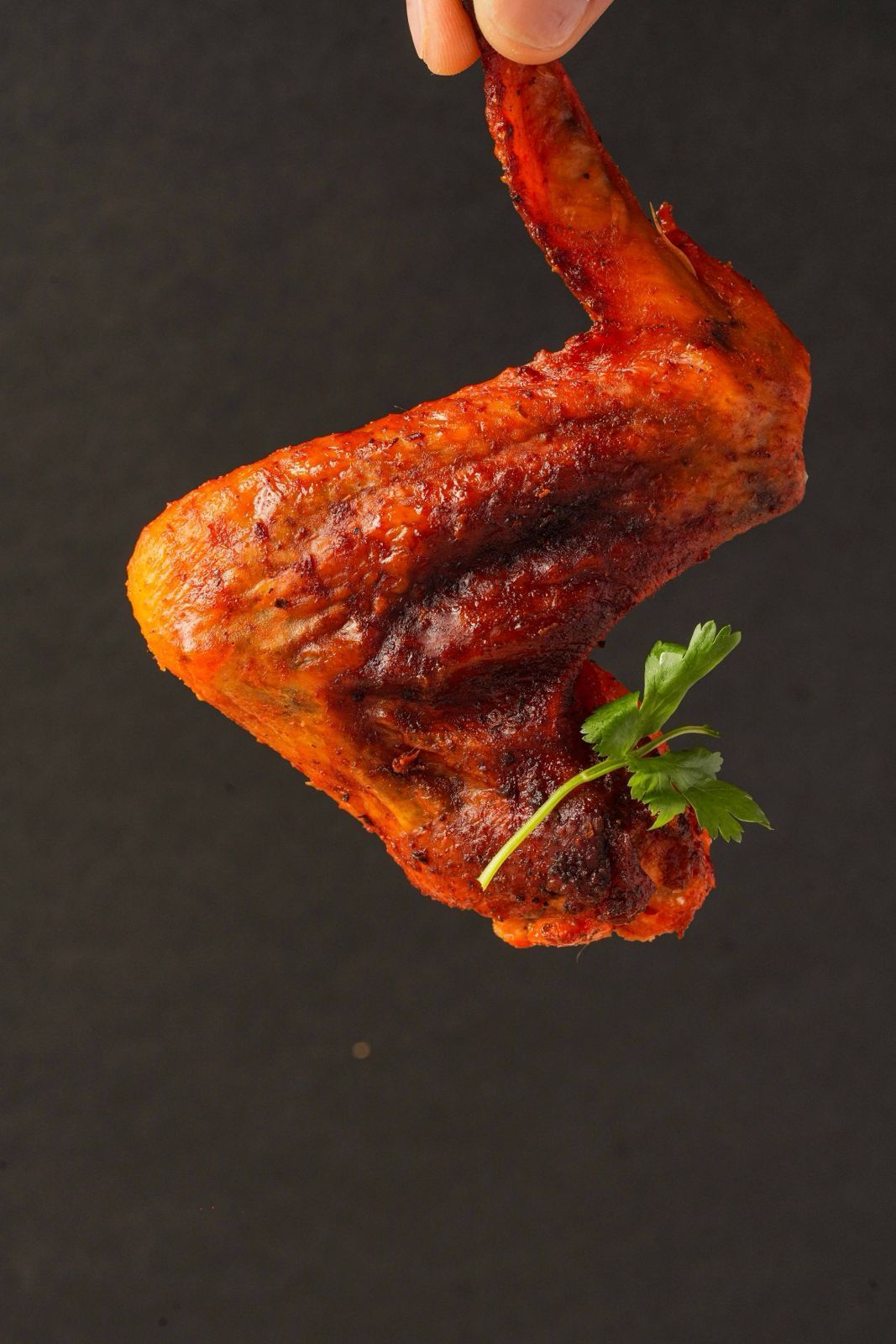 Crispy, golden-brown chicken wing held by a hand, with a sprig of parsley against a dark background.
