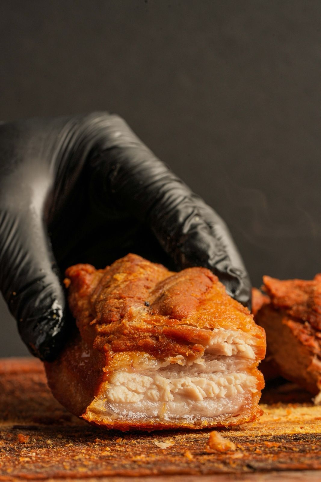 A gloved hand holds a piece of crispy pork belly, showing the tender, layered interior against a dark backdrop.