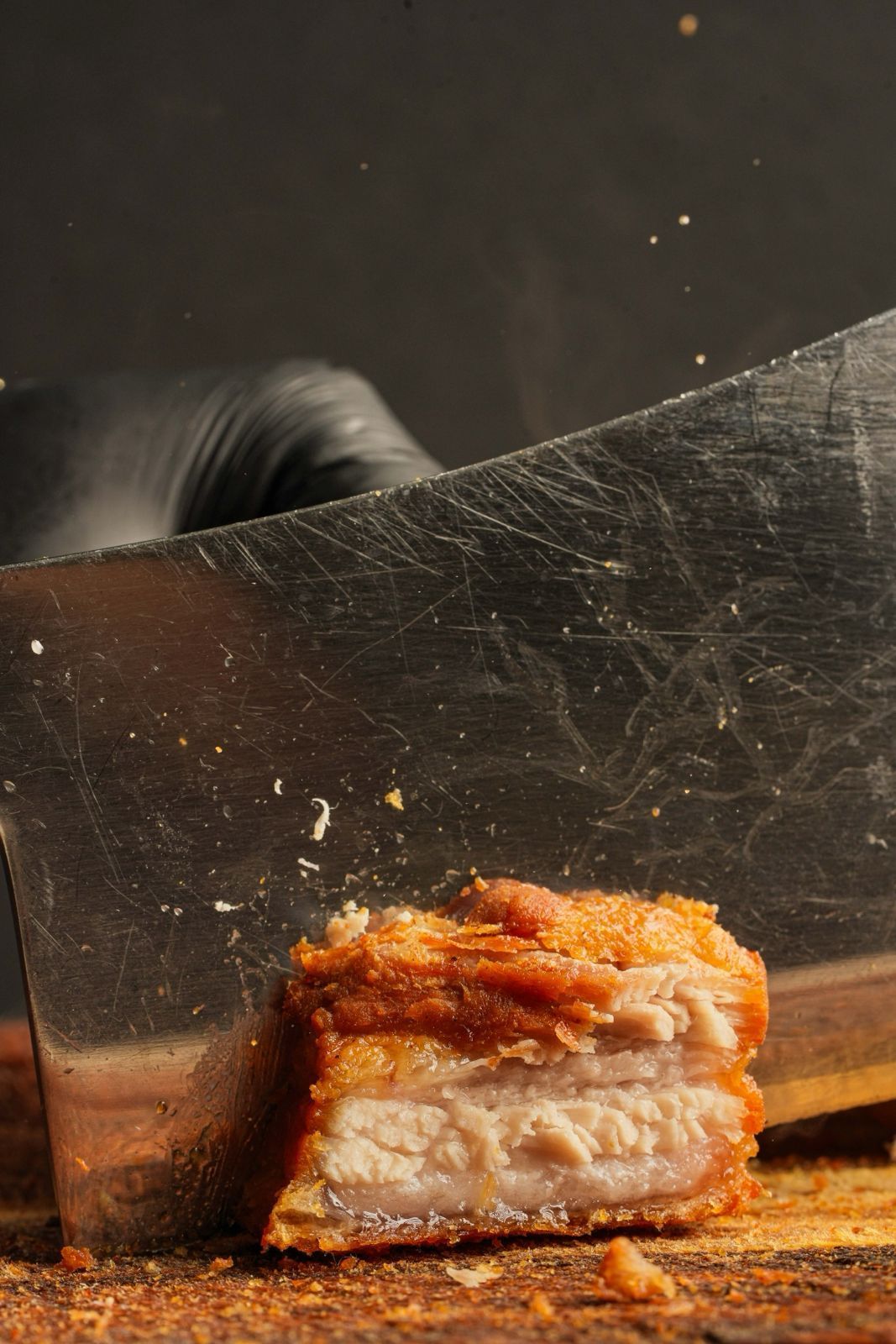 Cleaver slicing through a piece of crispy pork belly on a wooden board, bits of crackling flying.