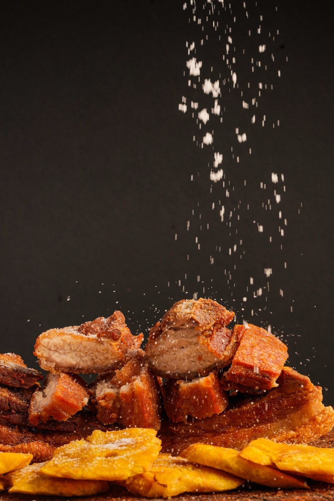 Fried pork belly and plantains with salt falling from above against a black background.
