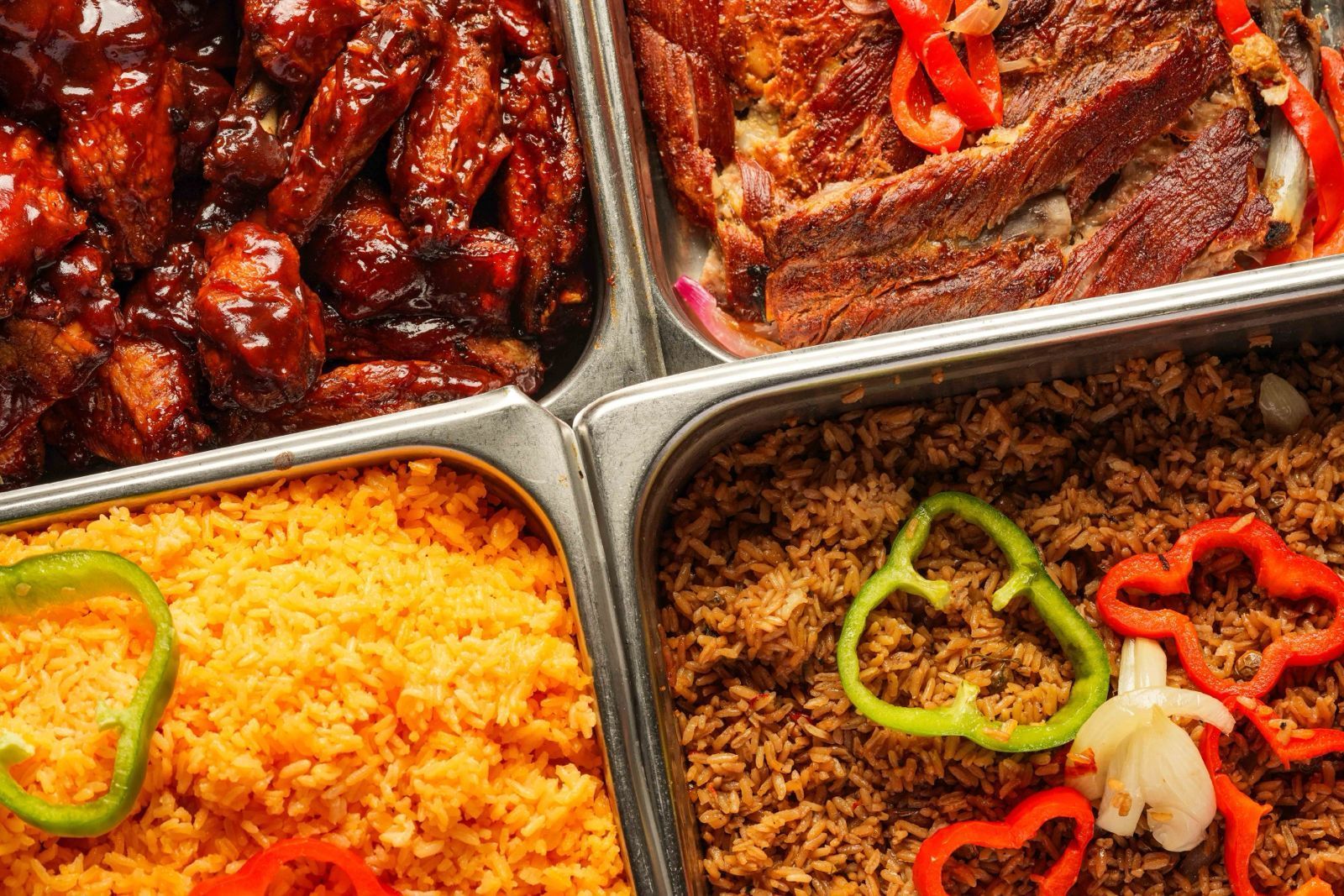 A buffet with four stainless steel trays holding: barbecue wings, roasted meat with vegetables, yellow rice,